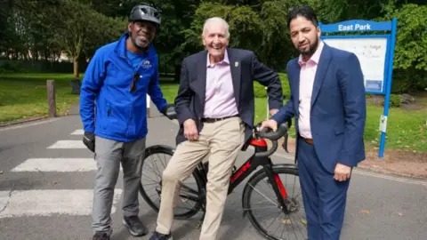 Three men, one seated on a stationary bike smile at the camera. 