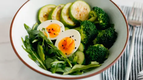 Getty Images A bowl with a hard boiled egg sliced in half, with salad leaves, broccoli and cucumber slices. 