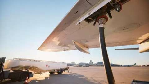 A close up of a plane wing with a jet fuel hose plugged in, and a fuel lorry in the background.