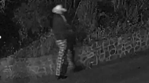 A black and white blurry screenshot from CCTV footage. It shows a man wearing a light coloured cowboy style hat, chequered trousers and a dark, long sleeved jumper. He is walking alongside a wall in the image.