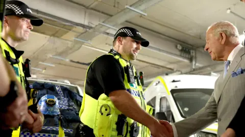 PA Media King Charles III in a light grey suit shakes hands with a police officer wearing a high-visibility vest inside what appears to be an emergency services facility. Several other uniformed officers stand nearby, with police equipment and a marked emergency vehicle visible in the background. Bright indoor lighting illuminates the industrial-style space, highlighting the uniforms, vehicle, and surrounding infrastructure.