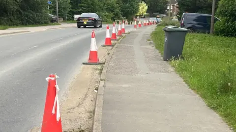 Graham Statter Line of red cones along a village street, with sporadic parked cars and a wheelie bin in view