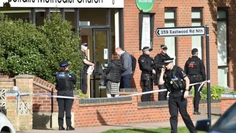 PA Media Armed police and members of the public outside a brick building in which MP Sir David Amess was killed in 2021