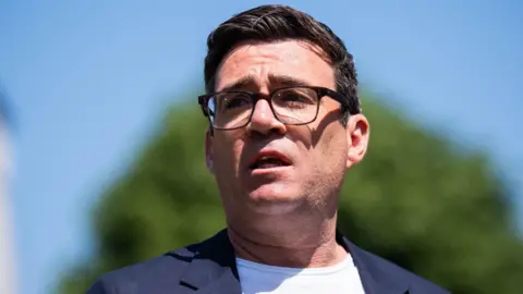 PA Andy Burnham wears a blue jacket and a white t-shirt. He has dark hair and wears glasses.