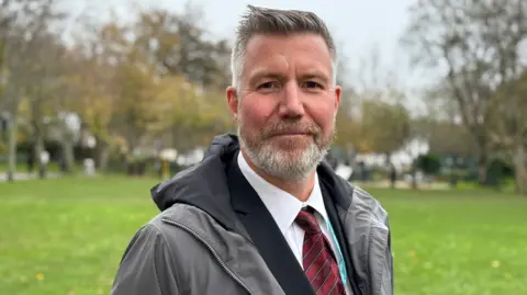 A man in his early 40s wearing a lack suit, white tie and red stripped tie. He also has a grey rain mac on and i stood in a park. He has a grey quiff and a grey beard.