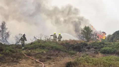 Tres bomberos del SFRS combaten un gran incendio en un páramo. Se pueden ver columnas de humo y llamas, así como una manguera enrollable.