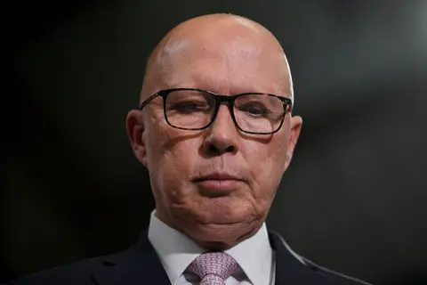 Peter Dutton, wearing glasses and a suit, looks down, his face neutral, in a close-up headshot