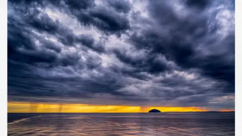 Craig Bradshaw Grey stormy skies mix with the yellow light of a sunset over an island