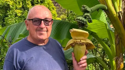 A head and shoulders image of a man wearing a T-shirt and glasses while holding a banana plant.