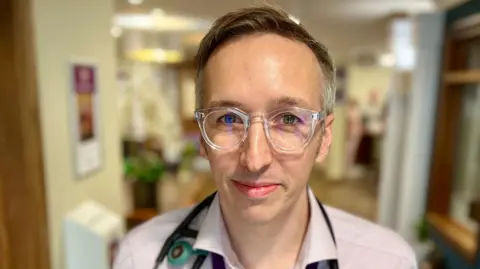 Matthew Butler has short brown hair and is wearing clear framed glasses, a pink shirt and has a stethoscope around his neck. He is standing in a hallway which has doors on either side and frames hung on the walls.