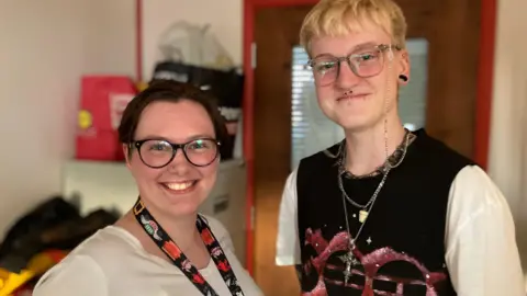 Jon Wright/BBC Katie Clark and Alex Fotheringham, stand side by side smiling at the camera. They are in a small office, with the background of the image slightly blurred.