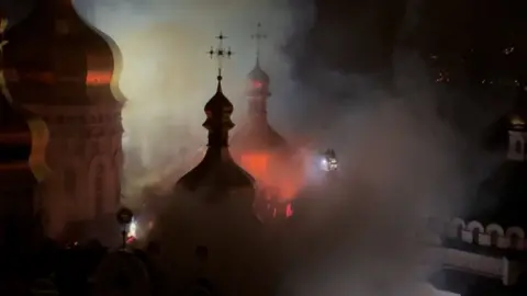 At night, fire glows on the cathedral roof as firefighters on a long ladder spray water on the site.