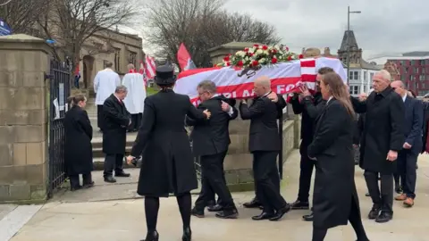 Men in black suits carry a coffin wrapped in Sunderland AFC's red and white flag. There is a bunch of red and white roses at its top. The procession is entering the ground of Sunderland Minster for a memorial service.