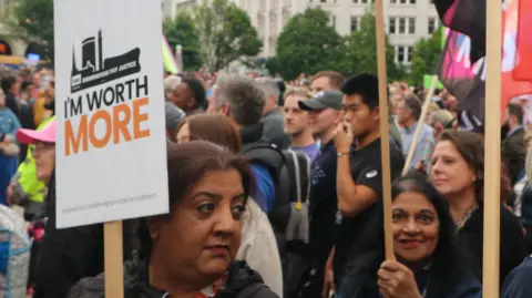 BBC Several women holding placards with one visible saying "I'm worth more" standing next to a crowd of people.