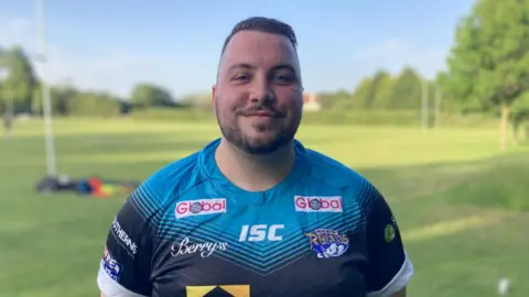 BBC/Sally Hurst Rugby player smiles at camera, with hands behind his back. He has a closely-cropped beard and moustache, and a nose stud.