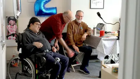 Three men, one of them in a wheelchair, smile as they sit in a room next to a table with a white tablecloth over it. They are playing a game that involves throwing an object a small distance.