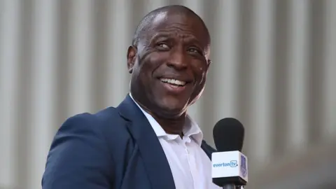 Kevin Campbell, wearing a dark navy suit jacket over a white shirt with an open collar, smiles while holding a microphone with the branding EvertonTV. 