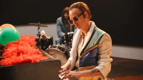 PA Media Cara Delevingne plays a brown piano dressed as Elton John. There are orange feathers and green and orange balloons on top of the piano. There is a drummer sat as a drum kit in the background.