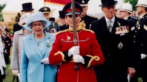 Manx National Heritage Queen Elizabeth II in a blue suit and hat with white gloves - standing behind someone in a red suit with white gloves holding a sword 