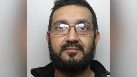 A police mugshot of Mohammed Khan. He has short dark hair and a beard. He is wearing glasses.