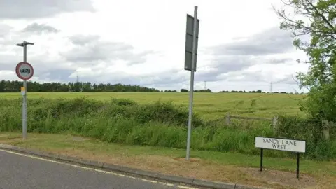 A view of a green field with a white sign reading Sandy Lane West.