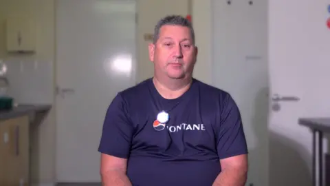 A man with short grey hair is facing the camera. He is wearing a navy t-shirt with white writing on it which can't quite be read. Behind him are white doors