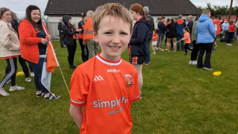 A young boy with blonde hair in an orange Armagh jersey smiles at the camera