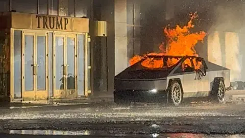 A Tesla Cybertruck with its windows blown out sits outside the Trump hotel on fire.