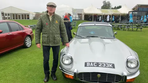 Anthony Evans, Classic Car Enthusiast talked about remembering the ring road when it first opened on voxes. He wears a green car and a green jacket and is next to a silver vintage sports car 