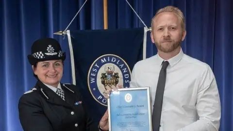 West Midlands Police A woman and a man hold a framed blue certificate. The woman is on the left in full police uniform. The man is on the right has brown-blonde hair and a brown beard and is wearing a white shirt and grey tie.