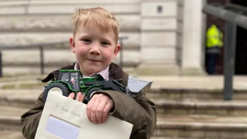 Malcolm Prior/BBC Five-year-old Bertie Church holding up a letter addressed to the Chancellor and a green toy tractor while standing outside the Treasury