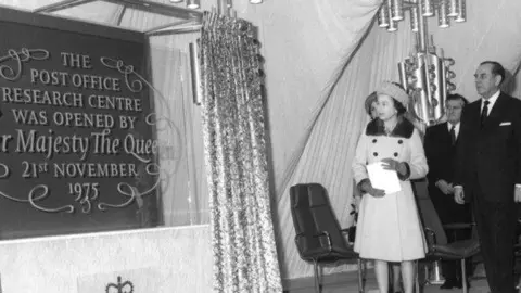 BT Group A black and white photo of Queen Elizabeth II unveiling a large plaque during the official opening of Adastral Park as it is now known. She wears a hat, gloves and dress, and is holding a piece of paper she looks at the plaque. Several men stand beside her wearing suits. 