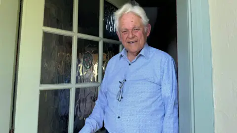 Pierre Tostevin standing in his doorway, he is wearing a blue shirt