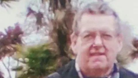 An older man standing in front of trees. He is wearing glasses, a coat and a scarf. 