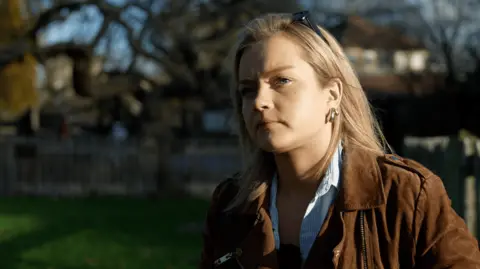 Georgia Poncia/BBC Young woman sitting down in a park. She has blonde hair and has sunglasses on top of her head. 