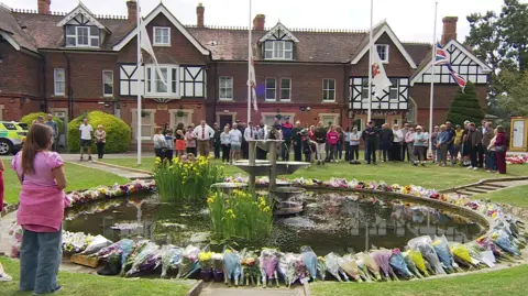 People gathered around a pond in Garth Park, observing a minute's silence. Flags behind a line of people are at half mast.