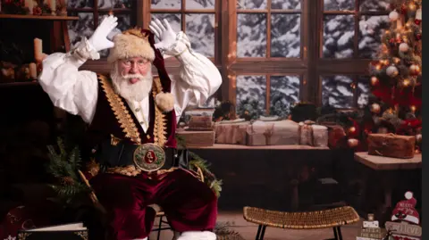 Danielle Axworthy Allan Evans dressed as Santa, sticking his tongue out with his hands in the air, in front of a snowy window background and a Christmas tree