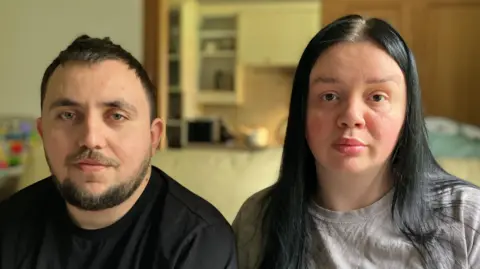 BBC / John Bowness Stepan and Alina Kozariichuk sit side by side in a living room and look directly at the camera with neutral expressions on their faces. On the left, Stepan is wearing a black shirt and has short black hair and a beard. Alina is wearing a grey shirt and has long black hair.
