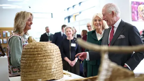 Reuters King Charles III and Britain's Queen Camilla meet willow artist Louise McLean during a visit to the Roe Valley Arts and Cultural Centre, in Limavady. Louise has blonde hair, wearing a crochet vest and light green shirt underneath. The king is pointing and smiling at the willow art work on the table.