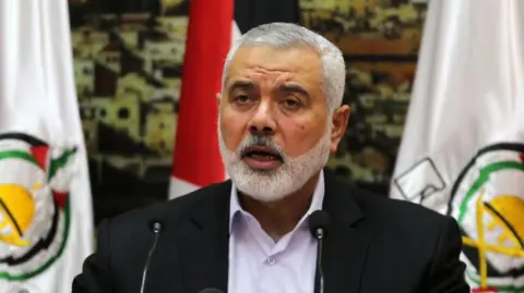 NurPhoto via Getty Images Ismael Haniyeh speaking into microphones with flags waving behind him