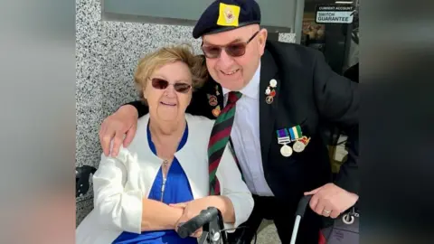 Roy and Marian Muir. Mrs Muir is sat down while Mr Muir has his arm around her and is crouching down. Mrs Muir is wearing a blue top with a white blazer, while Mr Muir is in a black suit, adorned with army medals. They both wear sunglasses.