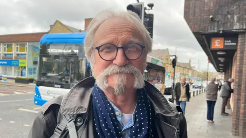 Trevor Burn is smiling slightly at the camera. He has a manicured, grey moustache and goatee and is wearing a blue pair of glasses. There is a single-decker bus on the road behind him and people using the Sainsbury's cash machine on his right.