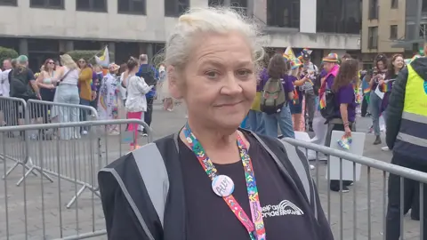 A woman is smiling. She's dresses in black and wearing a colourful lanyard. Crowds are behind her.