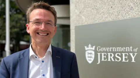 Kirsten is wearing glasses, a white shirt and a navy blazer, standing smiling outside a modern stone building next to a sign that reads “Government of Jersey.”