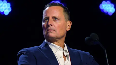 Richard Grenell, with short brown hair, wears a blue suit jacket and white shirt, as he stands in front of a microphone on the stage at a district meeting.