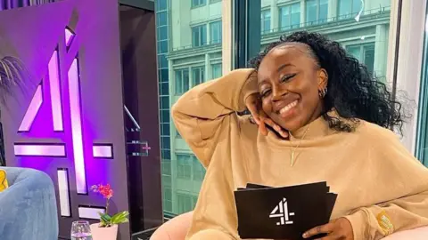 Stacey Olika Olika wearing a biscuit-beige lounge set, sitting on a pink chair on a Channel 4 set with a big number 4 neon sign to the left of her. She is smiling and has her long, curly black hair scraped back off her face and down her back.
There are big windows behind her, with buildings beyond. 