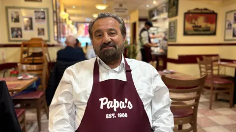 Adam sits against the backdrop of his restaurant, smiling and wearing a burgundy pinafore with Papa's EST. 1966 written across the front in white