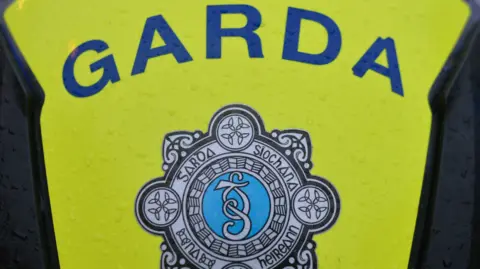 A close up of the emblem of the Garda Siochana on a motorbike. The word garda is printed across a neon yellow cover. It's been raining.