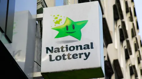 A white exterior sign hangs from a grey wall. The sign has a green star, with a black mouth and black eyes. Below the star are the words "National Lottery."