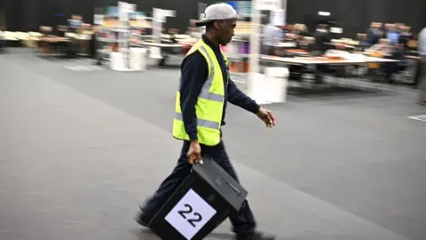 A man in a hi-vis and baseball cap worn backwards carries a ballot box with the number 22 on it. The background in the hall is out of focus.